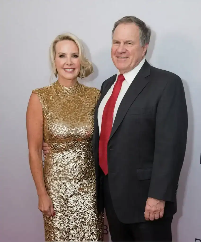 Former NFL coach Bill Belichick in a dark suit and red tie standing next to a smiling blonde woman in a sparkling gold dress on a red carpet.