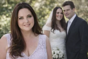 profit Paula smiling bridesmaid in a light purple lace dress with a bride and groom blurred in the background.